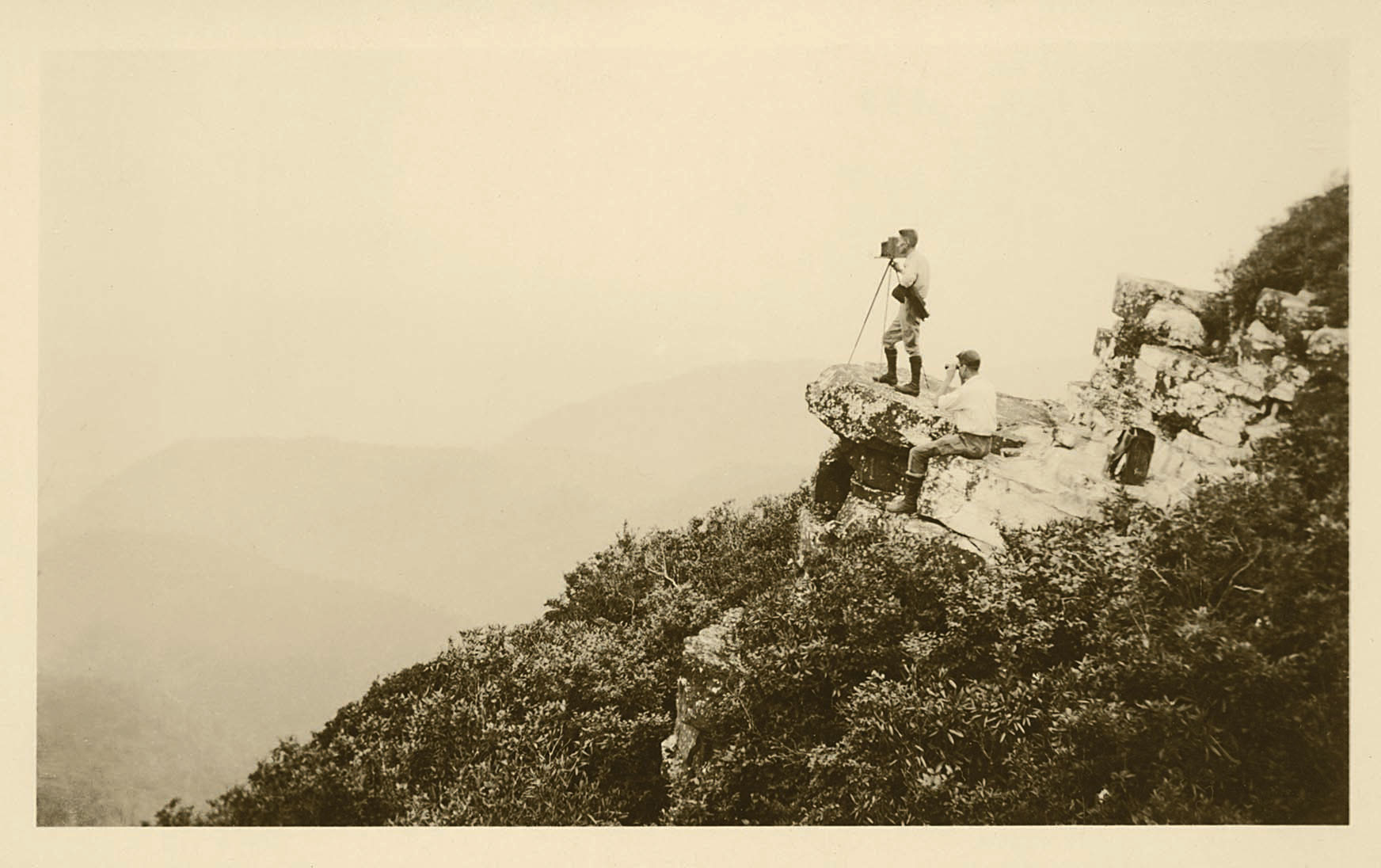 Black and white photo of two men in the distance standing on a rock with a camera