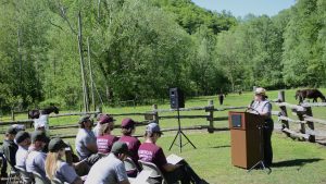 A woman in a ranger uniform stands at a podium on front of a seated crowd outside in a field.